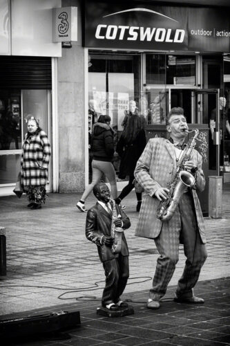 A man dressed in a 1950s-style suit, playing a saxophone. Next to him is a half-sized statue of another man playing the saxophone