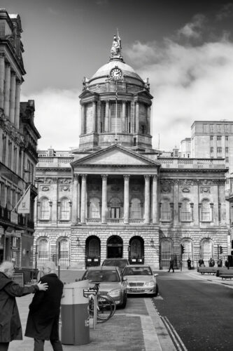 Looking at the front of the town hall. It's an 18th century building with two floors and a tall dome on top with a statue of Minerva. IN the foreground and two men talking