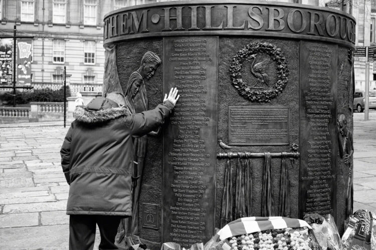 a man stands with his head bowed and his hand placed on the bronze memorial on the list of names of the deceased