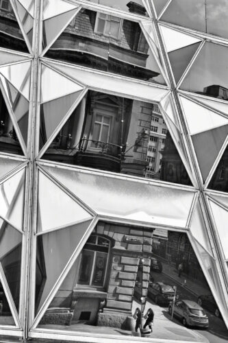 Geometric and angular glass on the side of a building. In the glass, a 19th century building is reflected. The reflection is a little disjointed and broken because of the angular glass