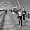 Coming towards the camera, which is looking down the pier, are two teenage girls on skateboards. One of the them is holding a balloon and looking at the floor, the other is looking at the camera