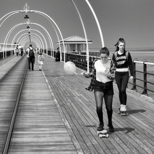 Coming towards the camera, which is looking down the pier, are two teenage girls on skateboards. One of the them is holding a balloon and looking at the floor, the other is looking at the camera