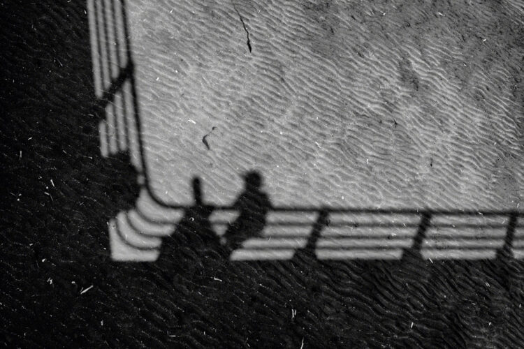 Looking down at the sand directly beneath the pier. The pier is casting a dark shadow on the sand, as a child standing on the pier and a telescope stand