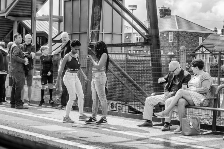 Two teenage girls on a train platform. One of them is dancing. There are other people standing nearby.