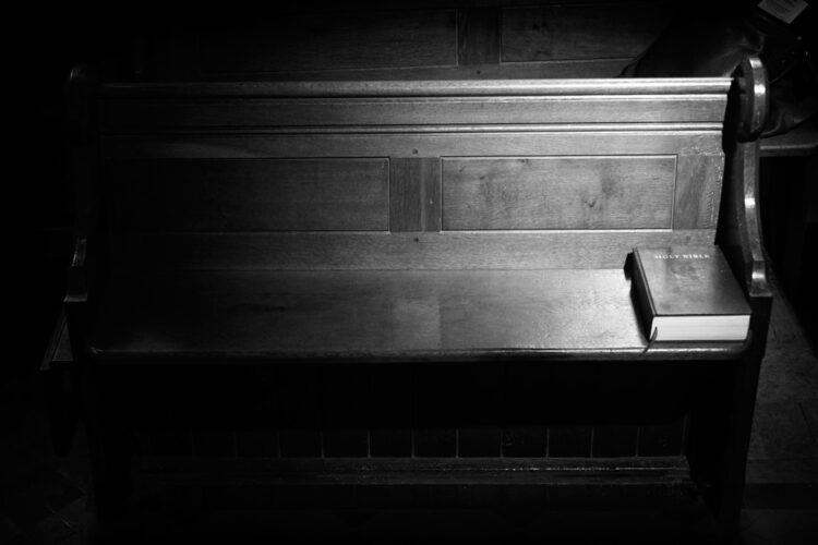 A wooden pew in partial darkness. In the part where the light is shining is a copy of the Bible