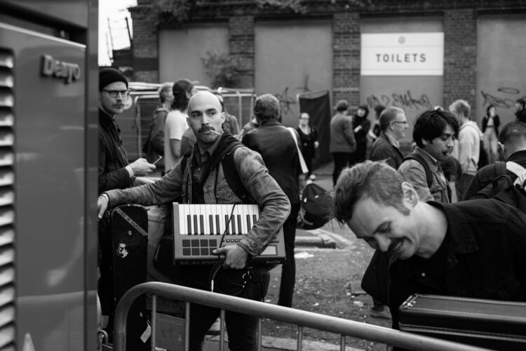 A man with a shaved head is carrying a small keyboard and is looking over his shoulder. Next to him is a man looking down at something and smiling. There are people around them as it's a music festival