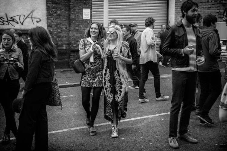 Two women walking down the street wearing sixties-style dresses. One has reflective sunglasses