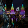 Looking across from first-floor level at the large Christmas tree made of hearts, lit up in rainbow colours. Shoppers are walking on the concourse in front of the tree