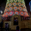 The two girls at the base of the large Christmas tree. The tree has small, wooden huts around its base, which have people sitting inside them.