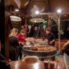 Looking at the interior of a circular, German-style bratwurst stall. There are three women all picking sausages off a round, central barbeque-type grill for customers.