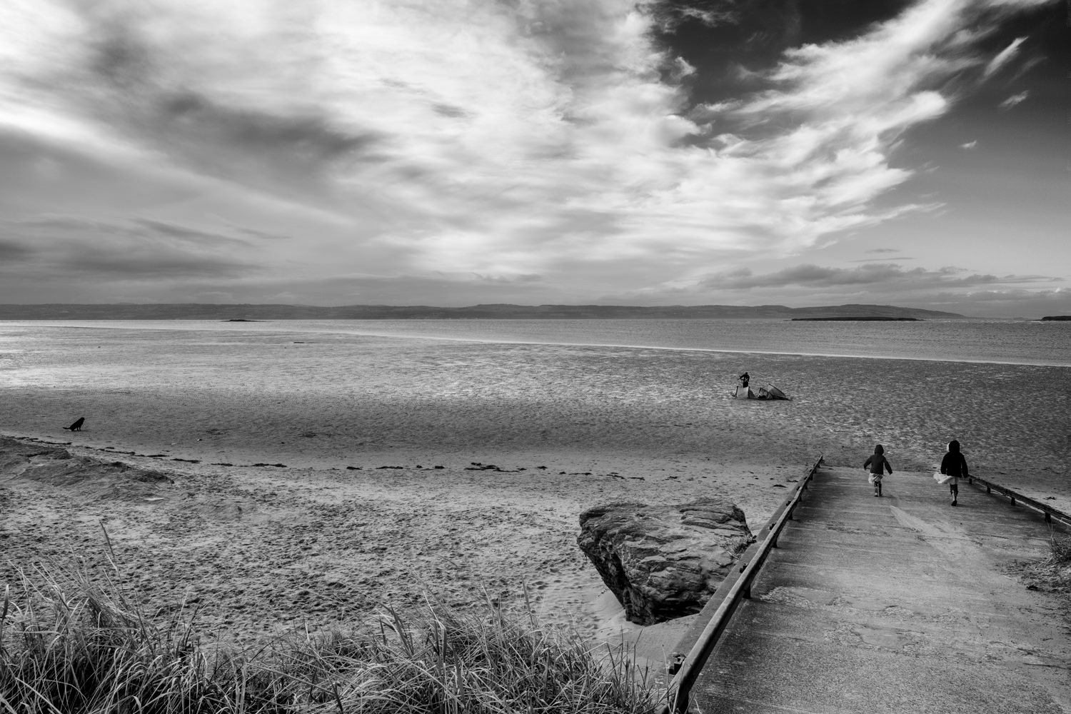 Looking across a sandy beach towards the sea. The North Wales coast is visible across the sea and the sky has dramatic clouds. There are two girls walking on to the beach