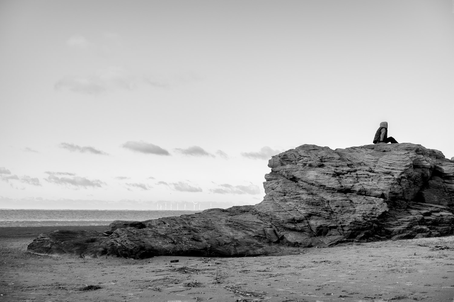 A boy sitting on top of a rocky outcrop, sitting side on to the camera. In the distance a wind farm can be seen out at sea