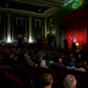 A woman on a stage dressed up as an old woman, talking to the audience