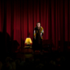 A woman on a stage dressed up as an old woman, talking to the audience