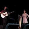 A man and woman on stage. The man is playing a guitar and the woman is singing