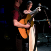 A man and woman on stage. The man is playing a guitar and the woman is smiling at the man