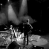 A man and woman on stage, seen from behind. The woman's looking at the man, who's playing a guitar. They have spotlights shining down on the them. There are a few members of the audience just visible