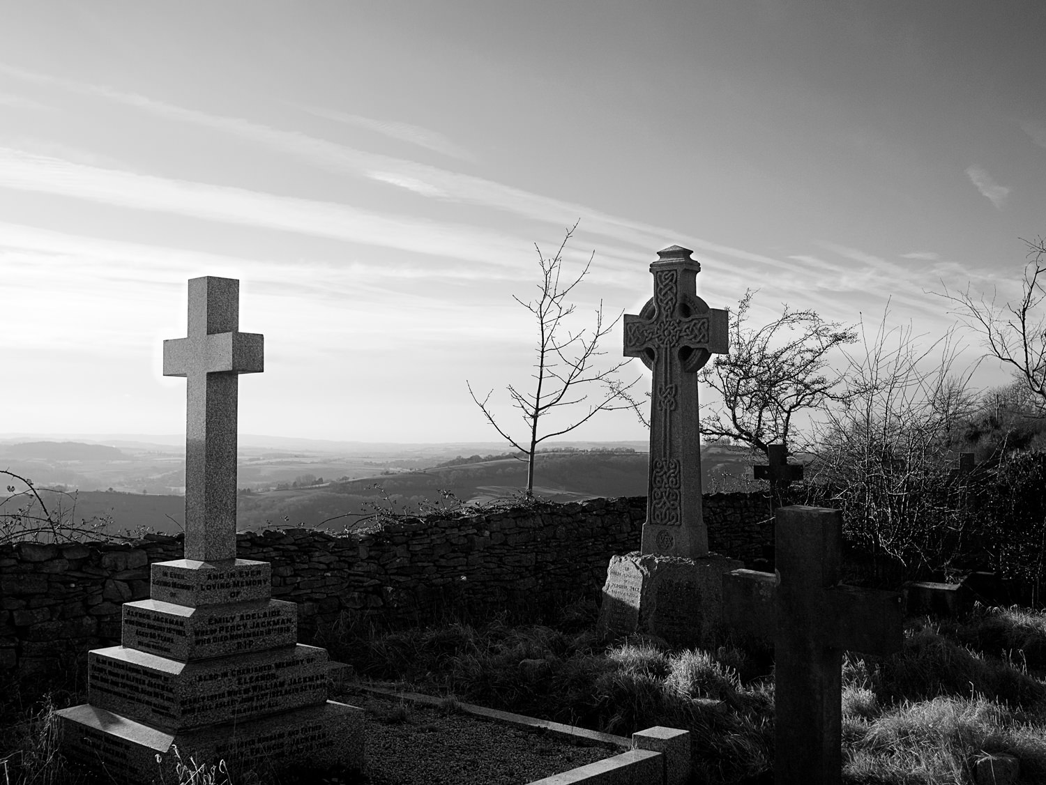 At the edge of Lansdown cemetery, looking over a low, stone wall, the valley stretches out as far as the eye can see. In the foreground are two graves with tall crosses. One cross is plain and the other is ornate, in the style of ancient Celtic crosses