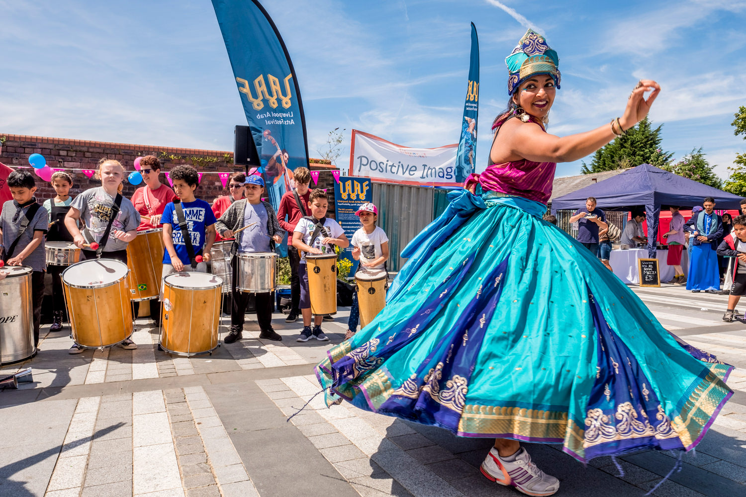 A woman in a traditional Indian-style dress dancing. Behind her is a troupe of drummers.