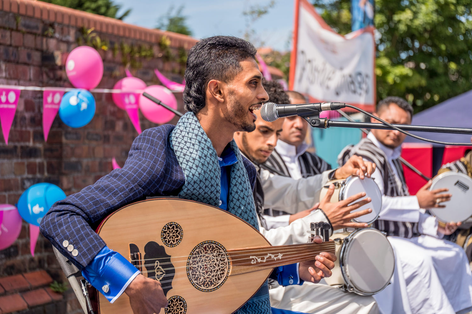 A traditional Arabic music group. Five seated men in front of a red-brick wall. Only the nearest man is in focus. He's singing into a microphone and playing an oud, a traditional Middle Eastern stringed instrument.