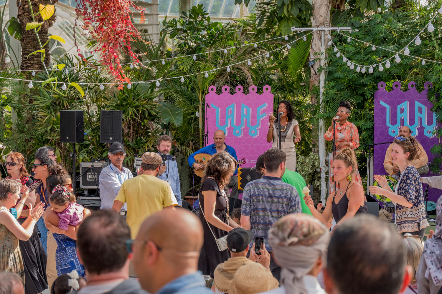 The group are performing on stage. In front is a group of people dancing, laughing and smiling.