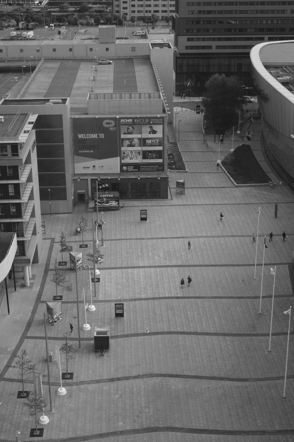 Looking down at a rectangular plaza in front of the Liverpool Echo Arena. Some people are walking along the plaza.